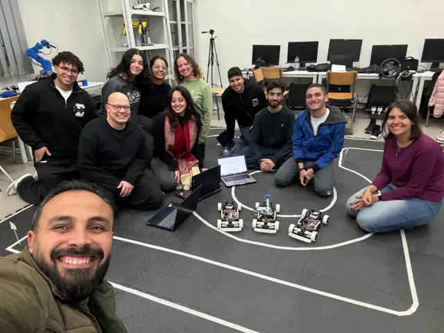 The RAVEN GP team and supervisors sitting around three small cars on a black foam-tile track.