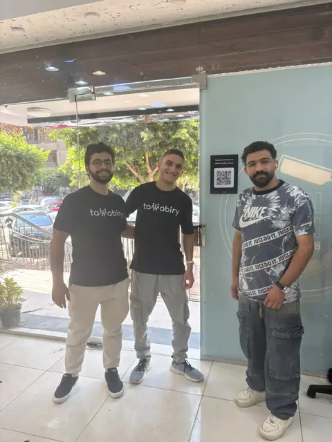 Two Tawabiry team members and the barbershop owner by the door, where the queue QR code is stuck.