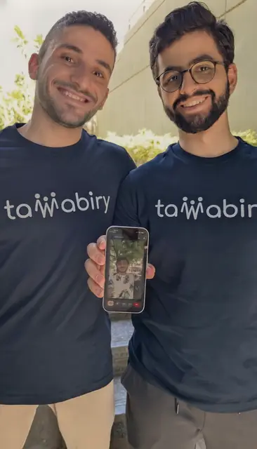 Two men in navy Tawabiry T-shirts holding up a phone on a video call.